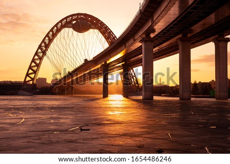 The photo shows picturesque bridge with big red arch over the river. This cable-stayed bridge stands on the frozen Moscow river. Crimson sunny rays illuminate bridge details and ice on the sunset.  Royalty-Free Stock Photo #1654486246