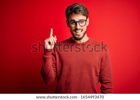 Young handsome man with beard wearing glasses and sweater standing over red background pointing finger up with successful idea. Exited and happy. Number one.