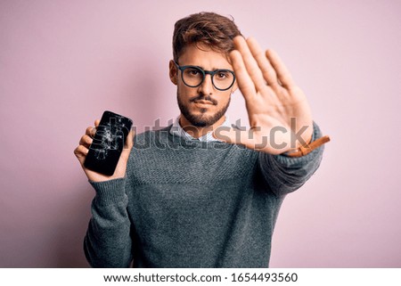 Young man with beard wearing glasses holding broken and craked smartphone with open hand doing stop sign with serious and confident expression, defense gesture