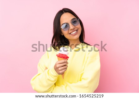 Young brunette girl holding a cornet ice cream over isolated pink background with glasses and smiling
