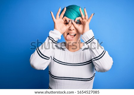 Young beautiful woman with blue fashion hair wearing casual sweater standing at studio doing ok gesture like binoculars sticking tongue out, eyes looking through fingers. Crazy expression.
