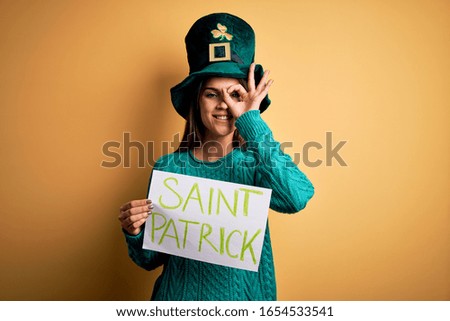 Woman wearing green hat celebrating st patricks day holding banner with saint patrick message with happy face smiling doing ok sign with hand on eye looking through fingers