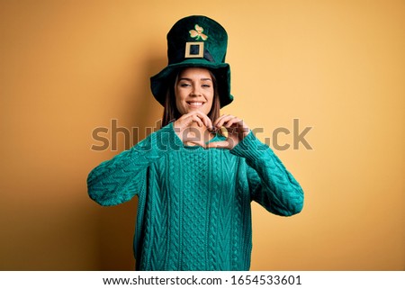 Young beautiful brunette woman wearing green hat with clover celebrating saint patricks day smiling in love doing heart symbol shape with hands. Romantic concept.