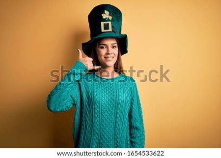 Young beautiful brunette woman wearing green hat with clover celebrating saint patricks day smiling doing phone gesture with hand and fingers like talking on the telephone. Communicating concepts.