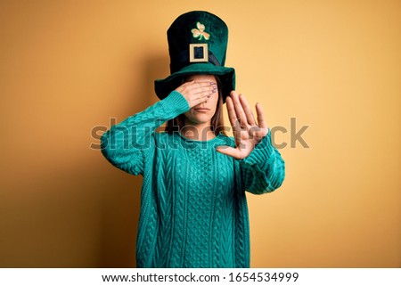 Young beautiful brunette woman wearing green hat with clover celebrating saint patricks day covering eyes with hands and doing stop gesture with sad and fear expression. Embarrassed and negative 