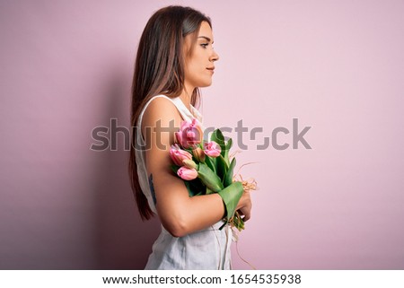 Young beautiful brunette woman holding bouquet of tulips flowers over pink background looking to side, relax profile pose with natural face and confident smile.