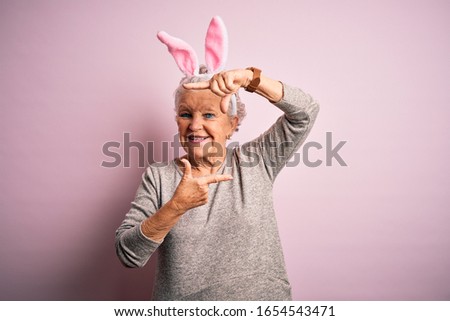 Senior beautiful woman wearing bunny ears standing over isolated pink background smiling making frame with hands and fingers with happy face. Creativity and photography concept.