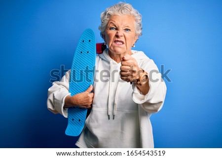 Senior beautiful sporty woman holding skate standing over isolated blue background annoyed and frustrated shouting with anger, crazy and yelling with raised hand, anger concept