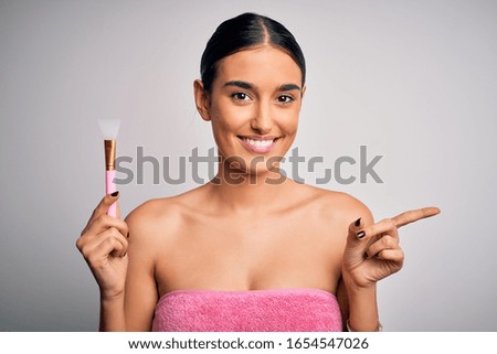 Young beautiful brunette woman wearing towel after shower applying cosmetics using spatula very happy pointing with hand and finger to the side