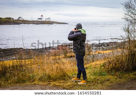 Young man with camera taking pictures of lighthouse by the sea far away in cold day
