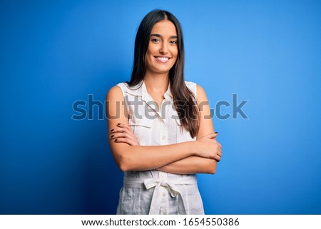 Young beautiful brunette woman wearing casual dress over isolated blue background happy face smiling with crossed arms looking at the camera. Positive person.