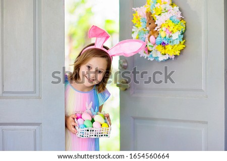Kids at Easter egg hunt. Child holding basket with colorful chocolate eggs, wearing bunny ears. Little girl at decorated front door after Easter celebration. Children celebrate spring holiday.