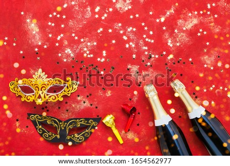 Two champagne bottles, carnival mask, tongue pipe and confetti on red background. Flat lay of Purim Carnival, New Year, Christmas celebration concept. Copy space, top view.