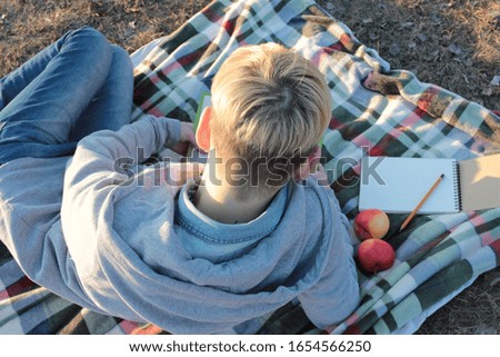 Young woman lying on a picnic blanket, reading a book in a park. Back view. Remote learning concept