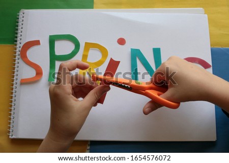 Scissors in the Hand and fingers of a child cut out different letters from bright paper for creativity. The word spring in colored letters on the table. Creative background with font symbols for kids 