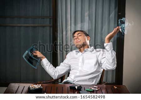 A guy with a picture of a lung X-ray, sitting at a table with an ashtray on it, lies a cigarette and a lighter with a cigarette in his mouth, leaning back in his chair. Humility with the consequences.