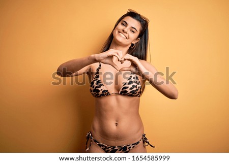 Young beautiful brunette woman on vacation wearing swimwear bikini over yellow background smiling in love doing heart symbol shape with hands. Romantic concept.