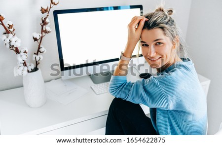 Smiling young freelance businesswoman using desktop pc in the office. The blonde female sits indoors at home working on a computer with a blank screen for your text message or content.