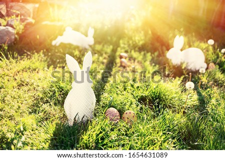 The concept of Easter holidays. Decorative Handicrafts handmade in the form of different white rabbits, located next to the painted eggs. Green grass in the background. Sunlight
