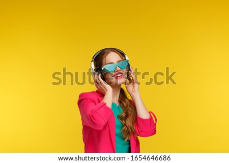 Enjoy listening to music. Young redhead woman in headphones listening music. Funny smiling girl in earphones and pink jacket dancing and singing on yellow background. Relaxation and stress management.