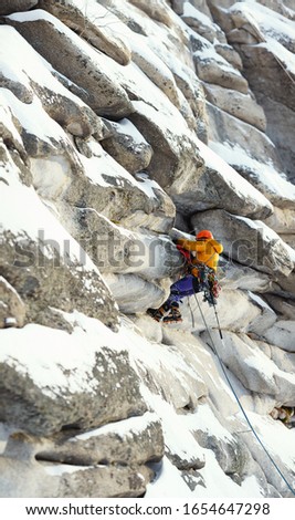 Mountaineer figure in bright outfit climbing up during the winter ascent in the mountains, against the background of overhanging rocks in the foreground and background are out of focus, panorama.