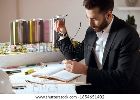 bearded good looking skilled architect making notes on notepad, close up side view photo.working time in the office.professional achitect developing interior and working on the planning of buildings.