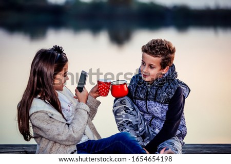 Two young cute little friends, boy and girl  having fun and drinking tea while sitting by the lake in the evening. Girl taking a photo with her phone of a cups of tea. Friendship