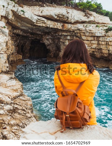 young woman sitting at cliff looking at grotto cave with blue azure water
