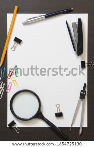 Template of white paper with pen, magnifier, stapler, paper clips and other stationery on dark wenge color wooden background. Stock photo with empty space for text 