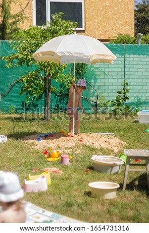 The boy holds an umbrella by the stock. A child sticks a beach umbrella in the ground. Children play in the yard on a sunny day.