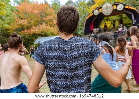 A twenty something boho guy is seen from the back, with unkempt brown hair and blue printed t shirt, dancing at festival with blurry people in background