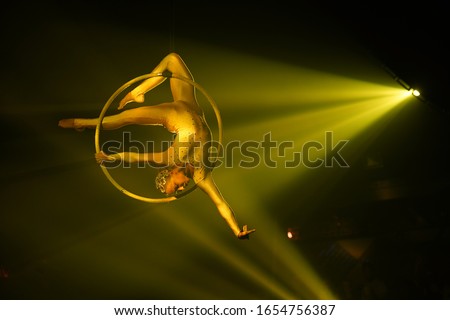 Flexible young woman make performance on aerial hoop, flexible back on aerial hoop, aerial circus show, yellow light. Flexible woman gymnast upside down on hoop. Golden costume, free space on right