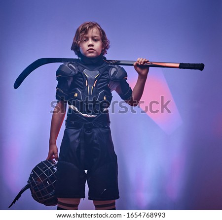 Young blonde, daring, sporty and strong boy, ice hockey player, posing in a bright neon studio for a photoshoot, wearing an ice-skating uniform while holding his helmet, hockey stick and standing in a
