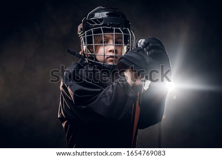 Young blonde sporty boy, ice hockey player, posing in a dark studio for a photoshoot, wearing an ice-skating uniform while wearing his helmet, holding hands on hockey stick and being quiet and calm