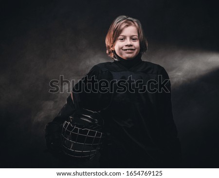 Young blonde sporty boy, ice hockey player, posing in a dark studio for a photoshoot, wearing an ice-skating uniform while holding his helmet and smiling