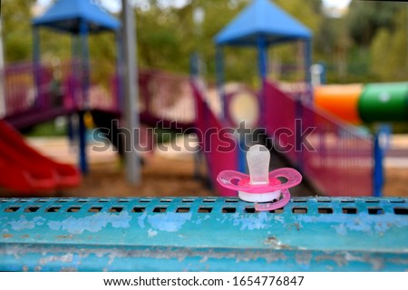 pink baby dummy (pacifier/soother) lost on the bench in an empty abandoned playground (park), with no children and without parents. cold, quiet and scary atmosphere.  Royalty-Free Stock Photo #1654776847