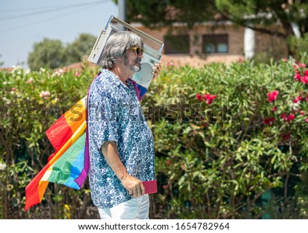 Gray-haired man with beard and moustache with sunglasses walking by a swimming pool with a cassette in the back, a mobile in the hand and a flag lgtb. Lifestyle