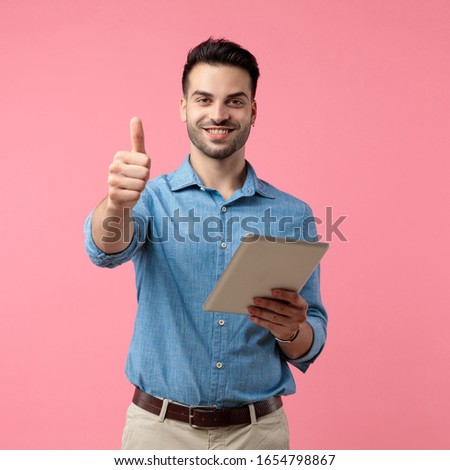 young casual guy making thumbs up sign and holding tab, standing on pink background 