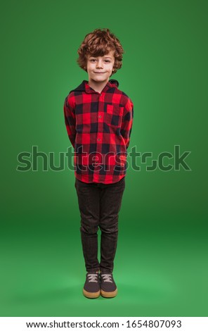 Full length adorable child in checkered shirt and jeans keeping hands behind back and looking at camera against green background