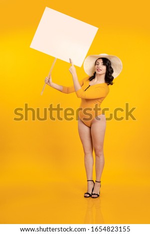 Young brunette girl with pin-up hairstyle in a yellow bathing suit and white hat holds a white poster in her hands and poses on a yellow background