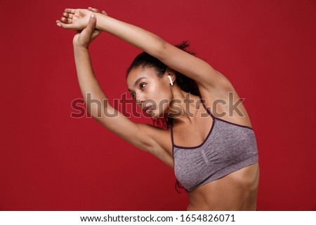 Beautiful african american sports fitness woman in sportswear working out isolated on red background. Sport exercises healthy lifestyle concept. Listen music with earpods, doing stretching exercising