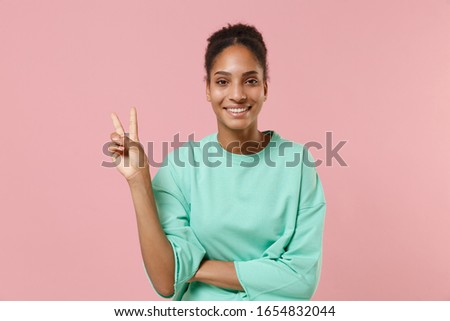 Cheerful young african american woman girl in green sweatshirt posing isolated on pastel pink background studio portrait. People emotions lifestyle concept. Mock up copy space. Showing victory sign