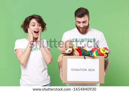 Amazed two friends couple in volunteer t-shirt isolated on green background. Voluntary free work assistance help charity grace teamwork concept. Hold donation box with kids toys, put hands on cheeks