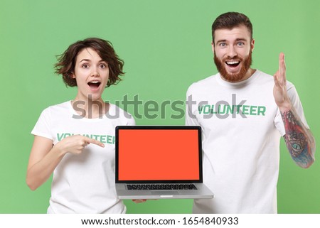 Excited friends couple in volunteer t-shirt isolated on green background. Voluntary free work assistance help charity grace teamwork. Point index finger on laptop pc computer with blank empty screen