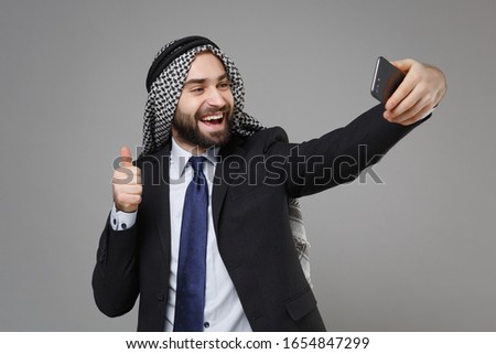 Cheerful arabian muslim businessman in keffiyeh kafiya ring igal agal suit isolated on gray background. Achievement career wealth business concept. Doing selfie shot on mobile phone showing thumb up