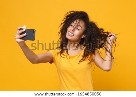 Cheerful young african american woman girl in casual t-shirt posing isolated on yellow orange background in studio. People lifestyle concept. Mock up copy space. Doing selfie shot on mobile phone