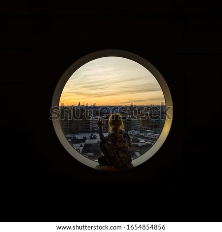 Young girl contemplating by looking through a round window during golden hour sunset.