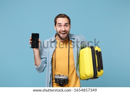 Pensive traveler tourist man in yellow clothes with photo camera isolated on blue background. Passenger traveling abroad on weekends. Air flight journey. Hold suitcase, mobile phone with blank screen.
