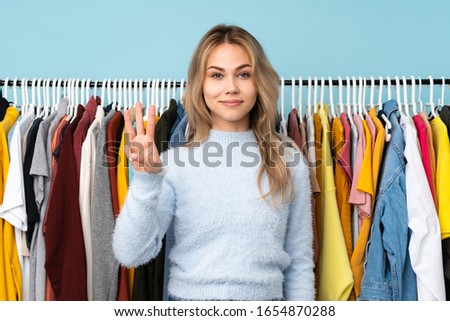 Teenager Russian girl buying some clothes isolated on blue background happy and counting three with fingers