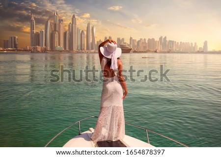 Woman in a white dress is standing on a boat looking to the skyline of Dubai Marina Royalty-Free Stock Photo #1654878397